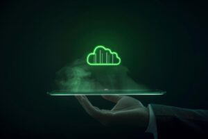 A imagem mostra uma mão segurando um dispositivo, possivelmente um tablet ou smartphone, do qual emerge um ícone digital de nuvem em neon verde, simbolizando tecnologia em nuvem (cloud computing). Há um efeito de luz e fumaça ao redor, destacando a inovação e a modernidade do conceito. O fundo escuro realça o contraste da nuvem luminosa, sugerindo um ambiente tecnológico e futurista.